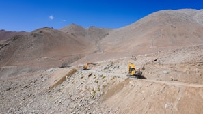 Heavy machinery operates on a rugged mountain landscape. Two yellow excavators are working on a dirt road that cuts through rocky terrain under a clear blue sky.