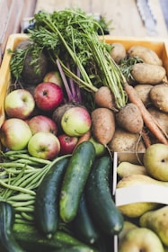 A wooden crate filled with a variety of fresh produce, including apples, cucumbers, green beans, carrots, potatoes, sweet potatoes, and pears. The vegetables and fruits are arranged close together, showcasing their vibrant colors and natural textures. The green tops of the carrots and the shiny skin of the cucumbers add to the freshness of the assortment.