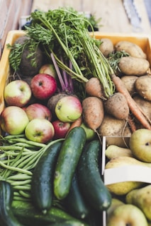 A wooden crate filled with a variety of fresh produce, including apples, cucumbers, green beans, carrots, potatoes, sweet potatoes, and pears. The vegetables and fruits are arranged close together, showcasing their vibrant colors and natural textures. The green tops of the carrots and the shiny skin of the cucumbers add to the freshness of the assortment.