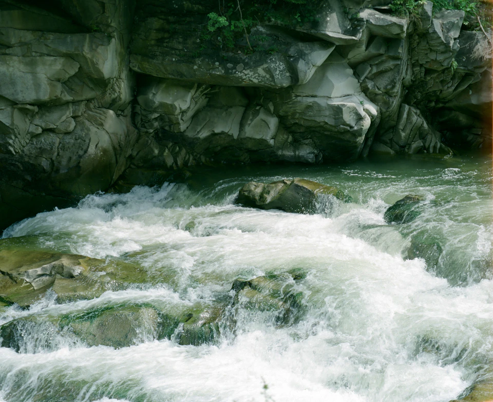 The image depicts a rocky river with white, frothy water flowing over large rocks and boulders. The river is surrounded by lush greenery, creating a serene and natural setting. The perspective of the photo is taken from above, providing an expansive view of the river's course and the surrounding landscape.