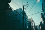 A cityscape featuring tall apartment buildings with multiple windows and external air conditioning units. Power lines crisscross against a clear blue sky, creating a network of wires leading to a central utility pole.