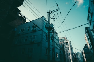 A cityscape featuring tall apartment buildings with multiple windows and external air conditioning units. Power lines crisscross against a clear blue sky, creating a network of wires leading to a central utility pole.