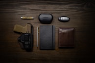 A neatly arranged flat lay of everyday carry items on a wooden surface. It includes a folding knife, a case labeled 'Bose', a car key fob, a firearm in a holster, a pen, a closed notebook, and a brown leather wallet.