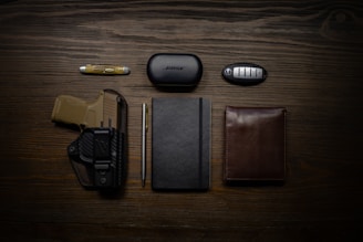 A neatly arranged flat lay of everyday carry items on a wooden surface. It includes a folding knife, a case labeled 'Bose', a car key fob, a firearm in a holster, a pen, a closed notebook, and a brown leather wallet.
