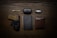 A neatly arranged flat lay of everyday carry items on a wooden surface. It includes a folding knife, a case labeled 'Bose', a car key fob, a firearm in a holster, a pen, a closed notebook, and a brown leather wallet.