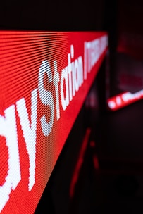 A close-up view of an LED display featuring bright red and white text. The text is prominently visible with a strong diagonal perspective, creating a dynamic visual effect. The background is blurred, emphasizing the sharpness of the LED lights.