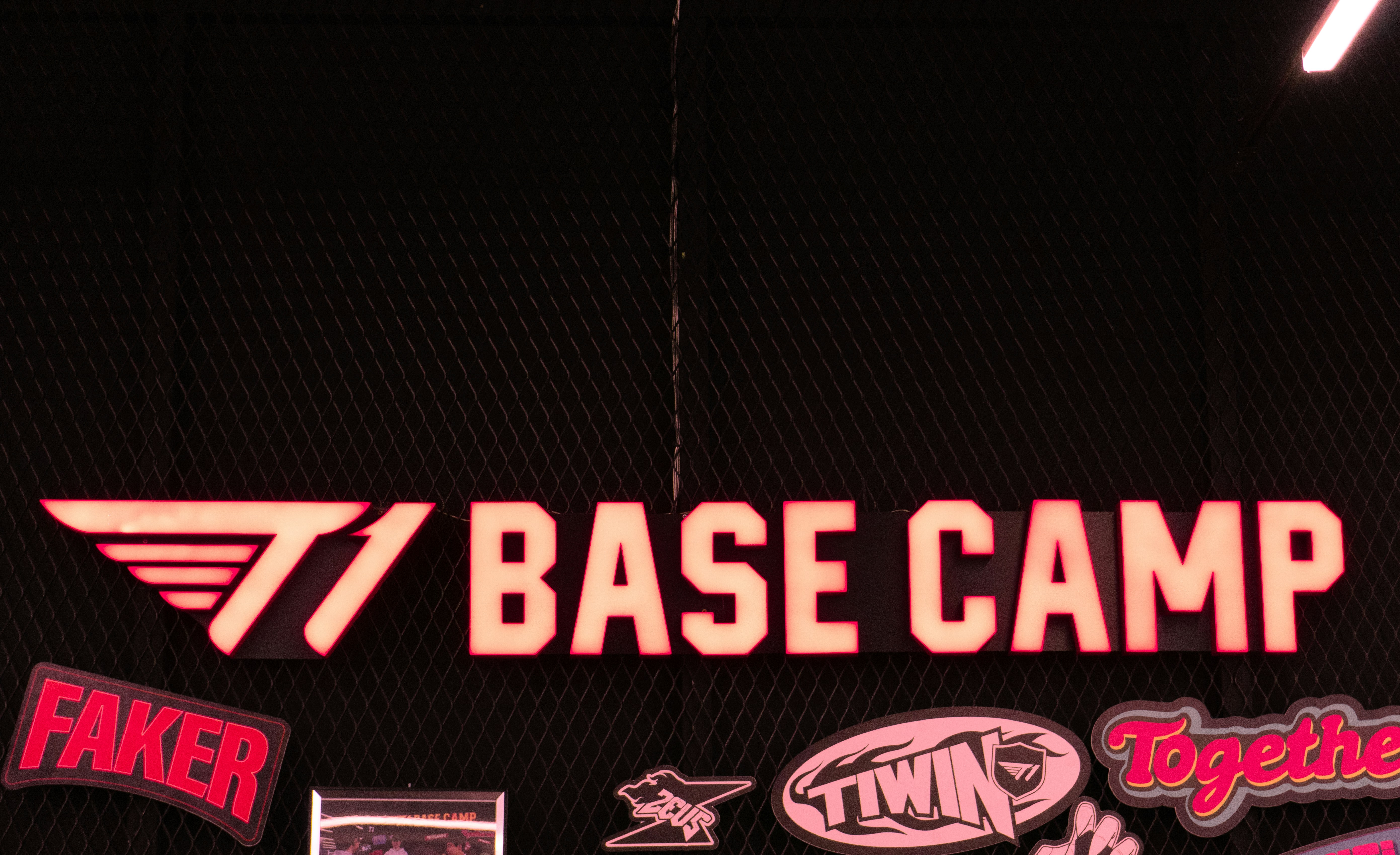A neon sign reading 'T1 BASE CAMP' is prominently displayed. The background features a dark mesh design. Below the main sign, there are various smaller logos and words such as 'FAKER', 'TWIN', and 'Together'. The overall design conveys a sense of team spirit and branding.