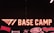 A neon sign reading 'T1 BASE CAMP' is prominently displayed. The background features a dark mesh design. Below the main sign, there are various smaller logos and words such as 'FAKER', 'TWIN', and 'Together'. The overall design conveys a sense of team spirit and branding.