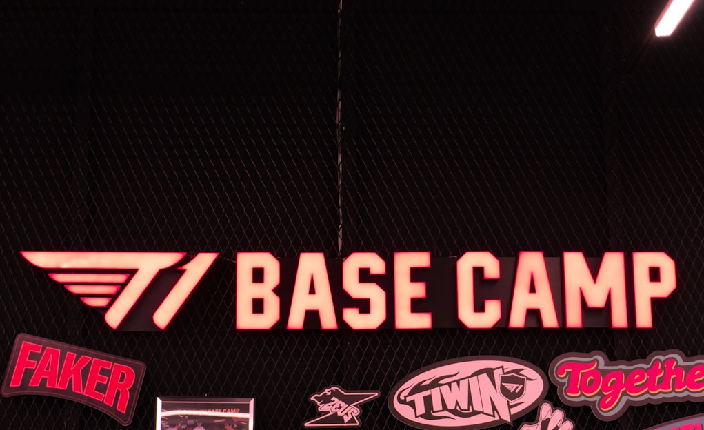 A neon sign reading 'T1 BASE CAMP' is prominently displayed. The background features a dark mesh design. Below the main sign, there are various smaller logos and words such as 'FAKER', 'TWIN', and 'Together'. The overall design conveys a sense of team spirit and branding.
