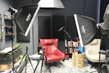A photography or recording studio setup featuring softbox lights, a red armchair, and a beige chair. There are tripods positioned around the chairs, and a microphone stand is visible. Books are neatly arranged on shelves attached to the wall, along with various decorative items. The room has a modern, organized feel with multiple lighting and recording equipment pieces.