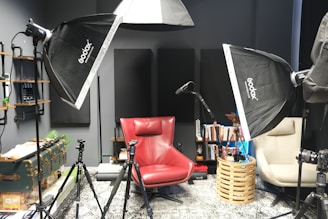 A photography or recording studio setup featuring softbox lights, a red armchair, and a beige chair. There are tripods positioned around the chairs, and a microphone stand is visible. Books are neatly arranged on shelves attached to the wall, along with various decorative items. The room has a modern, organized feel with multiple lighting and recording equipment pieces.
