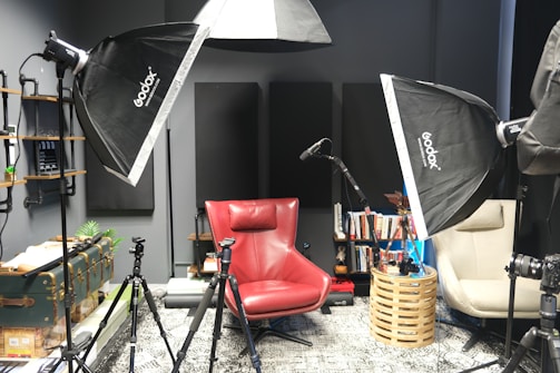 A photography or recording studio setup featuring softbox lights, a red armchair, and a beige chair. There are tripods positioned around the chairs, and a microphone stand is visible. Books are neatly arranged on shelves attached to the wall, along with various decorative items. The room has a modern, organized feel with multiple lighting and recording equipment pieces.