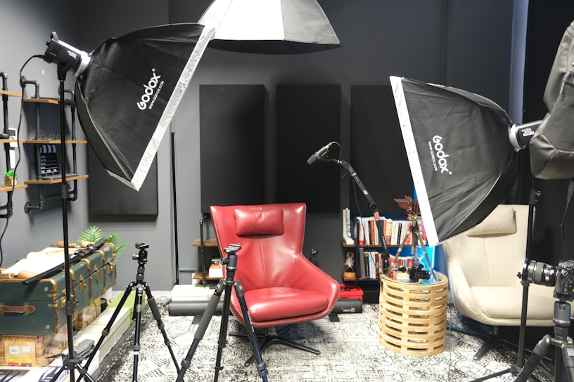 A photography or recording studio setup featuring softbox lights, a red armchair, and a beige chair. There are tripods positioned around the chairs, and a microphone stand is visible. Books are neatly arranged on shelves attached to the wall, along with various decorative items. The room has a modern, organized feel with multiple lighting and recording equipment pieces.