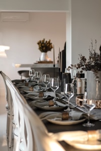 A long dining table is elegantly set with neatly folded napkins held by wooden rings, accompanied by wine glasses and plates. The table is decorated with minimalist candle holders and dried floral arrangements. In the background, a vase with flowers is placed on a table near a wall.
