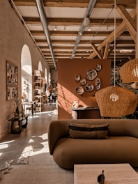 A cozy interior featuring a modern living area with earthy tones. A brown sofa is in the foreground, adorned with cushions. Wicker pendant lights hang from a ceiling with exposed wooden beams. Decorative plates are mounted on an orange-brown divider. Natural light streams through arched windows, illuminating the space. Plants and a wooden rack with various items enhance the warmth of the room.