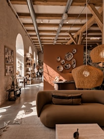 A cozy interior featuring a modern living area with earthy tones. A brown sofa is in the foreground, adorned with cushions. Wicker pendant lights hang from a ceiling with exposed wooden beams. Decorative plates are mounted on an orange-brown divider. Natural light streams through arched windows, illuminating the space. Plants and a wooden rack with various items enhance the warmth of the room.
