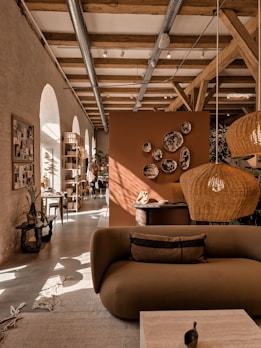 A cozy interior featuring a modern living area with earthy tones. A brown sofa is in the foreground, adorned with cushions. Wicker pendant lights hang from a ceiling with exposed wooden beams. Decorative plates are mounted on an orange-brown divider. Natural light streams through arched windows, illuminating the space. Plants and a wooden rack with various items enhance the warmth of the room.