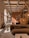 A cozy interior featuring a modern living area with earthy tones. A brown sofa is in the foreground, adorned with cushions. Wicker pendant lights hang from a ceiling with exposed wooden beams. Decorative plates are mounted on an orange-brown divider. Natural light streams through arched windows, illuminating the space. Plants and a wooden rack with various items enhance the warmth of the room.