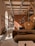 A cozy interior featuring a modern living area with earthy tones. A brown sofa is in the foreground, adorned with cushions. Wicker pendant lights hang from a ceiling with exposed wooden beams. Decorative plates are mounted on an orange-brown divider. Natural light streams through arched windows, illuminating the space. Plants and a wooden rack with various items enhance the warmth of the room.