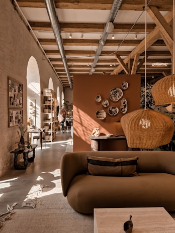 A cozy interior featuring a modern living area with earthy tones. A brown sofa is in the foreground, adorned with cushions. Wicker pendant lights hang from a ceiling with exposed wooden beams. Decorative plates are mounted on an orange-brown divider. Natural light streams through arched windows, illuminating the space. Plants and a wooden rack with various items enhance the warmth of the room.