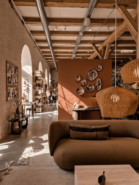 A cozy interior featuring a modern living area with earthy tones. A brown sofa is in the foreground, adorned with cushions. Wicker pendant lights hang from a ceiling with exposed wooden beams. Decorative plates are mounted on an orange-brown divider. Natural light streams through arched windows, illuminating the space. Plants and a wooden rack with various items enhance the warmth of the room.