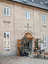 An old brick building with several white-framed windows and a dark gray tiled roof. The entrance has a wooden door surrounded by potted plants and small trees. In front of the entrance, there are outdoor tables and chairs, with a person sitting and reading or using a mobile device. Signage with the word 'ferm' is visible on the building.