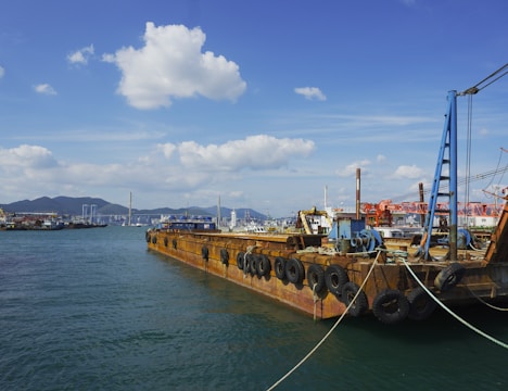 A large industrial barge is docked at a harbor, with numerous tires attached to its sides. The background includes a skyline with buildings and cranes, and a range of hills under a clear blue sky with scattered clouds.