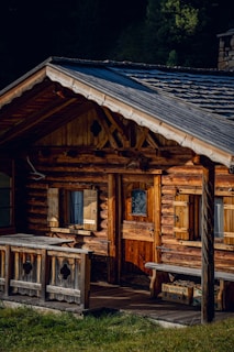 A rustic wooden cabin with a sloped roof and log construction is surrounded by lush greenery. The building features wooden shutters on the windows, a small wooden porch, and a wooden door with decorative ironwork. Sunlight highlights the natural textures and colors of the wood.