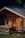 A rustic wooden cabin with a sloped roof and log construction is surrounded by lush greenery. The building features wooden shutters on the windows, a small wooden porch, and a wooden door with decorative ironwork. Sunlight highlights the natural textures and colors of the wood.