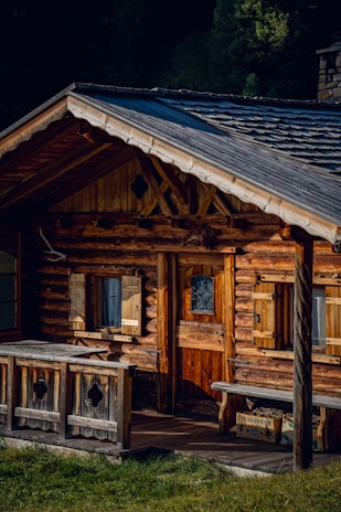 A rustic wooden cabin with a sloped roof and log construction is surrounded by lush greenery. The building features wooden shutters on the windows, a small wooden porch, and a wooden door with decorative ironwork. Sunlight highlights the natural textures and colors of the wood.