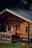A rustic wooden cabin with a sloped roof and log construction is surrounded by lush greenery. The building features wooden shutters on the windows, a small wooden porch, and a wooden door with decorative ironwork. Sunlight highlights the natural textures and colors of the wood.