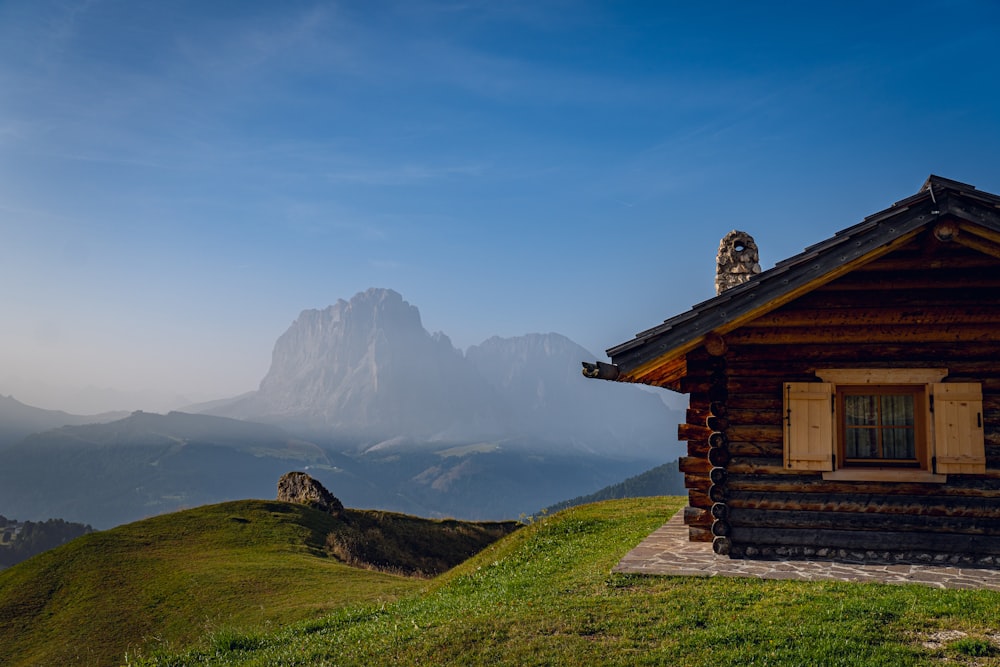 uma cabana de madeira em uma colina gramada com montanhas ao fundo