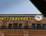 A building facade features a prominent clock with red hands and numbers, next to large, bold yellow letters spelling out 'AFTONBLADET.' The architecture includes decorative stonework and metal railings, and the sky appears clear.