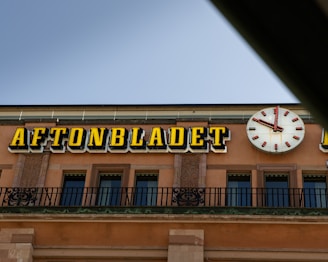A building facade features a prominent clock with red hands and numbers, next to large, bold yellow letters spelling out 'AFTONBLADET.' The architecture includes decorative stonework and metal railings, and the sky appears clear.