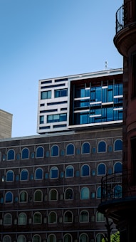 A modern building with a unique architectural design, featuring sections of arched windows and a contrasting top section with a grid of horizontal and vertical lines. The facade is composed of varying materials and colors, creating a striking visual contrast against the clear blue sky. Adjacent buildings with different styles are partially visible.