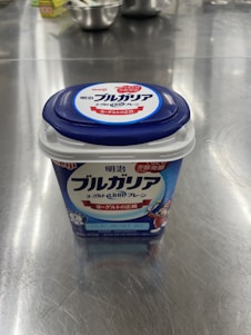 A container of Meiji Bulgarian yogurt is placed on a reflective metal surface. The packaging is predominantly blue with white and red accents. Several steel kitchen bowls and a box are blurred in the background.