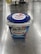 A container of Meiji Bulgarian yogurt is placed on a reflective metal surface. The packaging is predominantly blue with white and red accents. Several steel kitchen bowls and a box are blurred in the background.