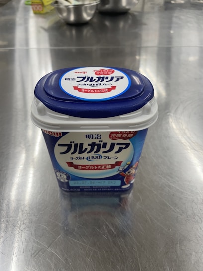 A container of Meiji Bulgarian yogurt is placed on a reflective metal surface. The packaging is predominantly blue with white and red accents. Several steel kitchen bowls and a box are blurred in the background.