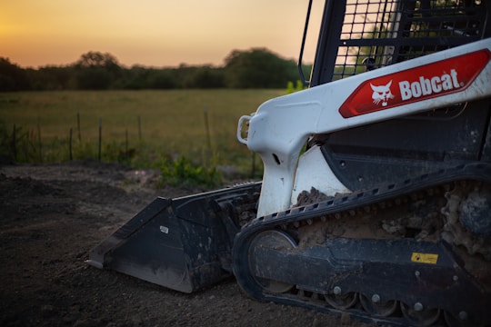 A close-up of a Bobcat skid steer loader with visible rubber tracks and part of the bucket attachment, positioned on dirt ground with a field and trees in the background under a clear sky with a warm glow.