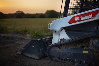 A close-up of a Bobcat skid steer loader with visible rubber tracks and part of the bucket attachment, positioned on dirt ground with a field and trees in the background under a clear sky with a warm glow.