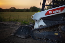 A close-up of a Bobcat skid steer loader with visible rubber tracks and part of the bucket attachment, positioned on dirt ground with a field and trees in the background under a clear sky with a warm glow.
