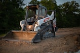 A compact track loader with a front-mounted bucket is parked on a dirt construction site. The machine has a white body with a logo on the side and wide tracks allowing it to move on uneven surfaces. Surrounding the vehicle are piles of dirt, and in the background, there are trees under a grayish sky.