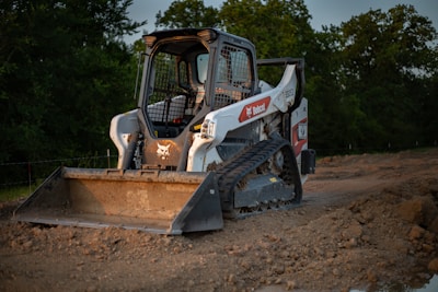 A compact track loader with a front-mounted bucket is parked on a dirt construction site. The machine has a white body with a logo on the side and wide tracks allowing it to move on uneven surfaces. Surrounding the vehicle are piles of dirt, and in the background, there are trees under a grayish sky.