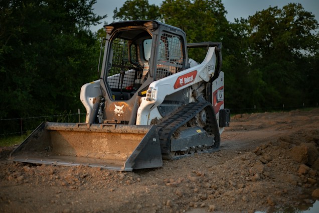 A compact track loader with a front-mounted bucket is parked on a dirt construction site. The machine has a white body with a logo on the side and wide tracks allowing it to move on uneven surfaces. Surrounding the vehicle are piles of dirt, and in the background, there are trees under a grayish sky.