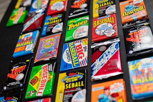 A collection of various vintage baseball card packs arranged on a dark surface. The packs feature vibrant and colorful designs with bold text, representing different brands and styles of baseball cards from past decades.