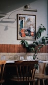 An interior space features a wooden dining table with chairs, set against a wall decorated with a framed painting and some green foliage. The wall is two-toned, with a section of red brick and a section of textured off-white paint. Above the painting is a classic lamp casting a shadow.
