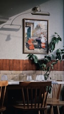 An interior space features a wooden dining table with chairs, set against a wall decorated with a framed painting and some green foliage. The wall is two-toned, with a section of red brick and a section of textured off-white paint. Above the painting is a classic lamp casting a shadow.