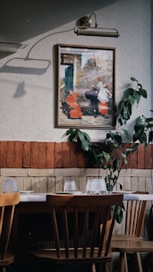 An interior space features a wooden dining table with chairs, set against a wall decorated with a framed painting and some green foliage. The wall is two-toned, with a section of red brick and a section of textured off-white paint. Above the painting is a classic lamp casting a shadow.
