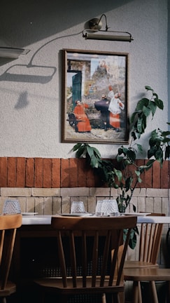 An interior space features a wooden dining table with chairs, set against a wall decorated with a framed painting and some green foliage. The wall is two-toned, with a section of red brick and a section of textured off-white paint. Above the painting is a classic lamp casting a shadow.