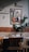 An interior space features a wooden dining table with chairs, set against a wall decorated with a framed painting and some green foliage. The wall is two-toned, with a section of red brick and a section of textured off-white paint. Above the painting is a classic lamp casting a shadow.